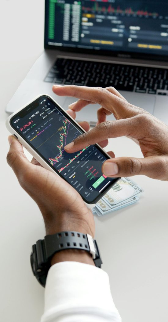 Hands using smartphone trading app with charts, dollar bills in background.