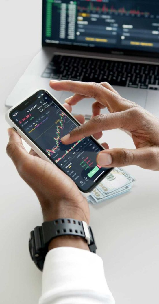 Hands using smartphone trading app with charts, dollar bills in background.